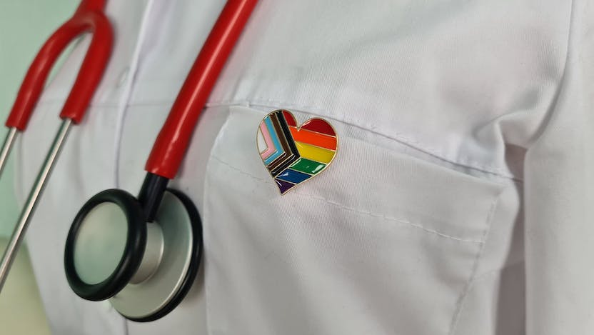 Close-up photograph of doctor's white coat with stethoscope and heart pin featuring LGBTQ pride flag