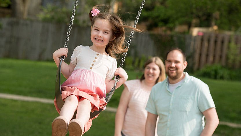 Joy Arseneau shown on an outdoor swing with her parents in the background.
